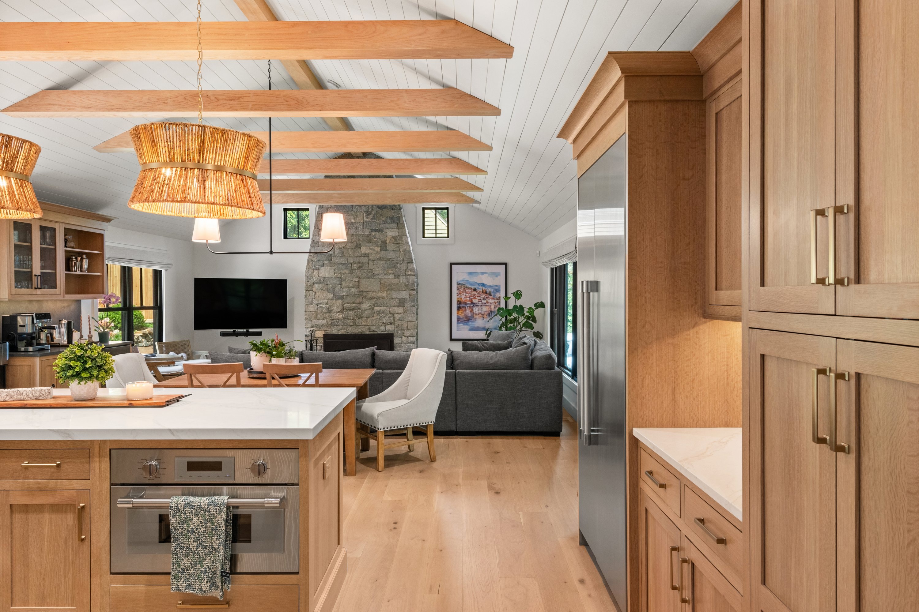 Open-plan kitchen and living room with oak cabinetry, exposed beams, shiplap ceiling, and stone fireplace