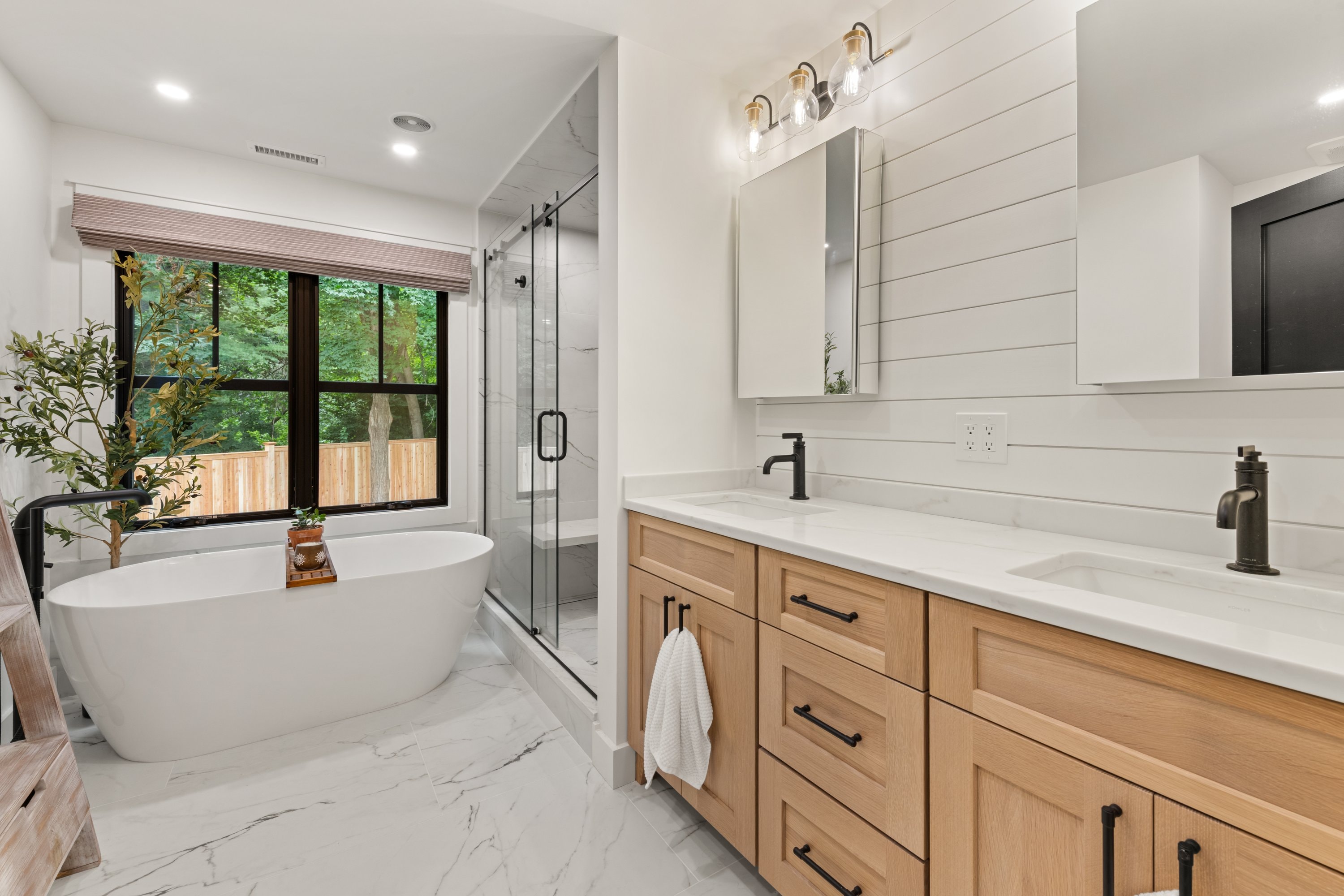 Primary bathroom with freestanding soaking tub, double oak vanity, marble tile, and black hardware